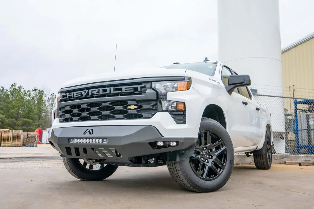 Fab Fours Vengeance front bumper on a white Chevrolet Silverado 1500, front driver-side three-quarter view outdoors, showing the center LED light bar, cube light pods, and slotted skid plate