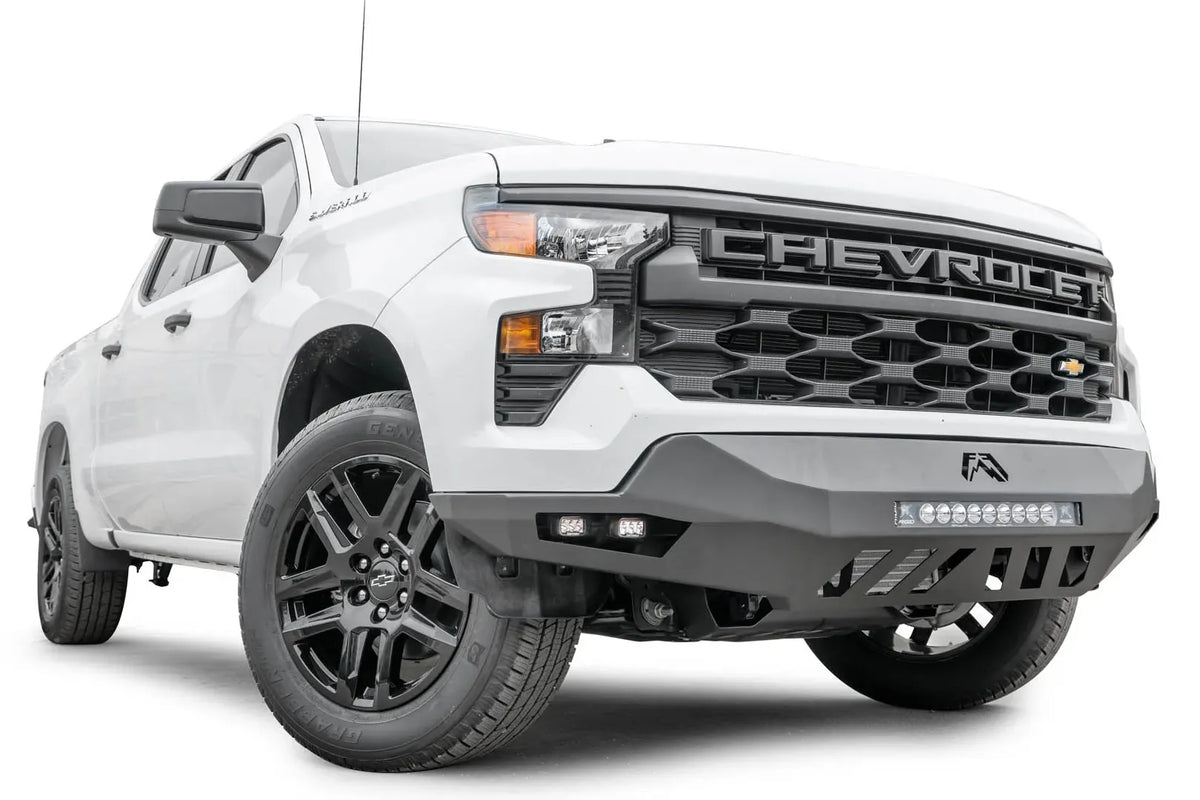 Fab Fours Vengeance front bumper on a white Chevrolet Silverado 1500, front passenger-side three-quarter view, showing the LED light bar, cube light pods, and slotted skid plate