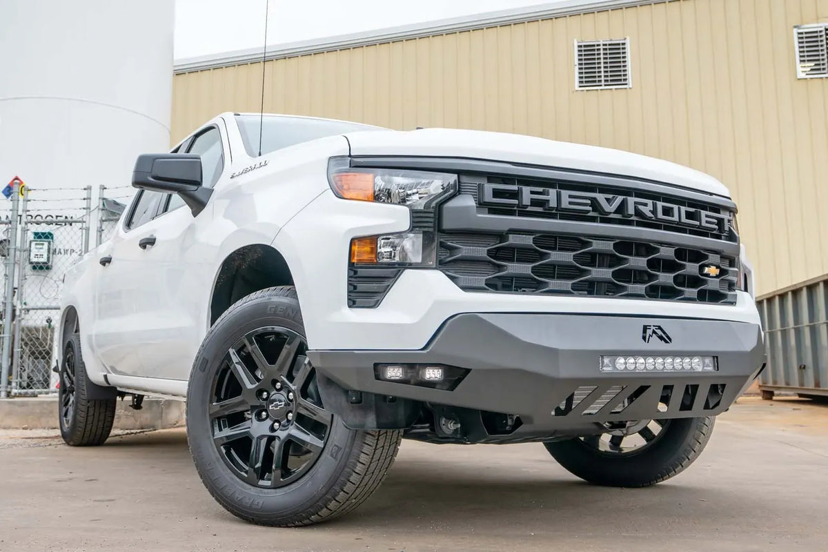 Fab Fours Vengeance front bumper on a white Chevrolet Silverado 1500, front passenger-side three-quarter view outdoors, showing the LED light bar, cube light pods, and skid plate