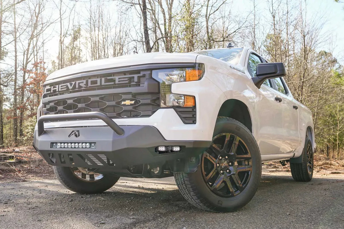 Fab Fours Vengeance front bumper with pre-runner guard on a white Chevrolet Silverado 1500, front driver-side three-quarter view outdoors, showing the tubular pre-runner bar, LED light bar, and cube light pods