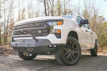 Fab Fours Vengeance front bumper with pre-runner guard on a white Chevrolet Silverado 1500, front driver-side three-quarter view outdoors, showing the tubular pre-runner bar, LED light bar, and cube light pods