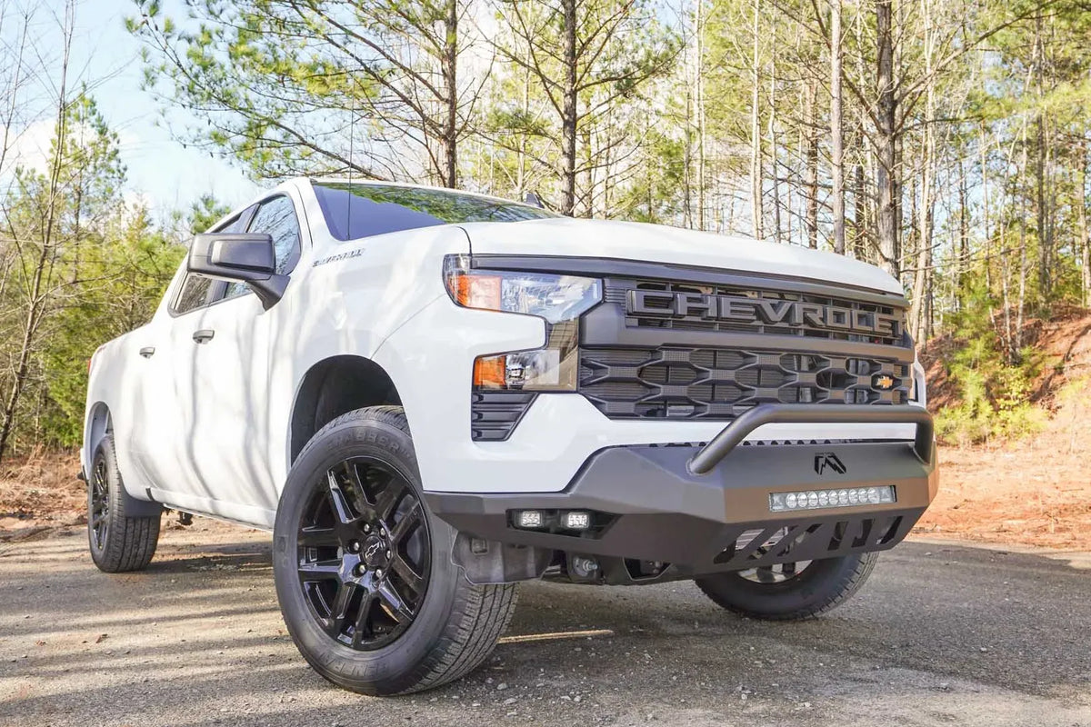 Fab Fours Vengeance front bumper with pre-runner guard on a white Chevrolet Silverado 1500, front passenger-side three-quarter view outdoors, showing the pre-runner bar, LED light bar, and angular bumper wing