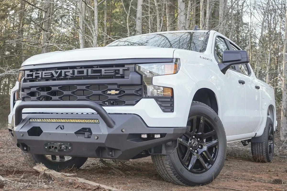 Fab Fours Matrix front bumper with pre-runner guard on a white Chevrolet Silverado 1500, low driver-side front three-quarter view in the woods, with LED light bar, cube lights, and winch fairlead