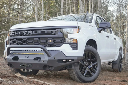 Fab Fours Matrix front bumper with pre-runner guard on a white Chevrolet Silverado 1500, low driver-side front three-quarter view in the woods, with LED light bar, cube lights, and winch fairlead