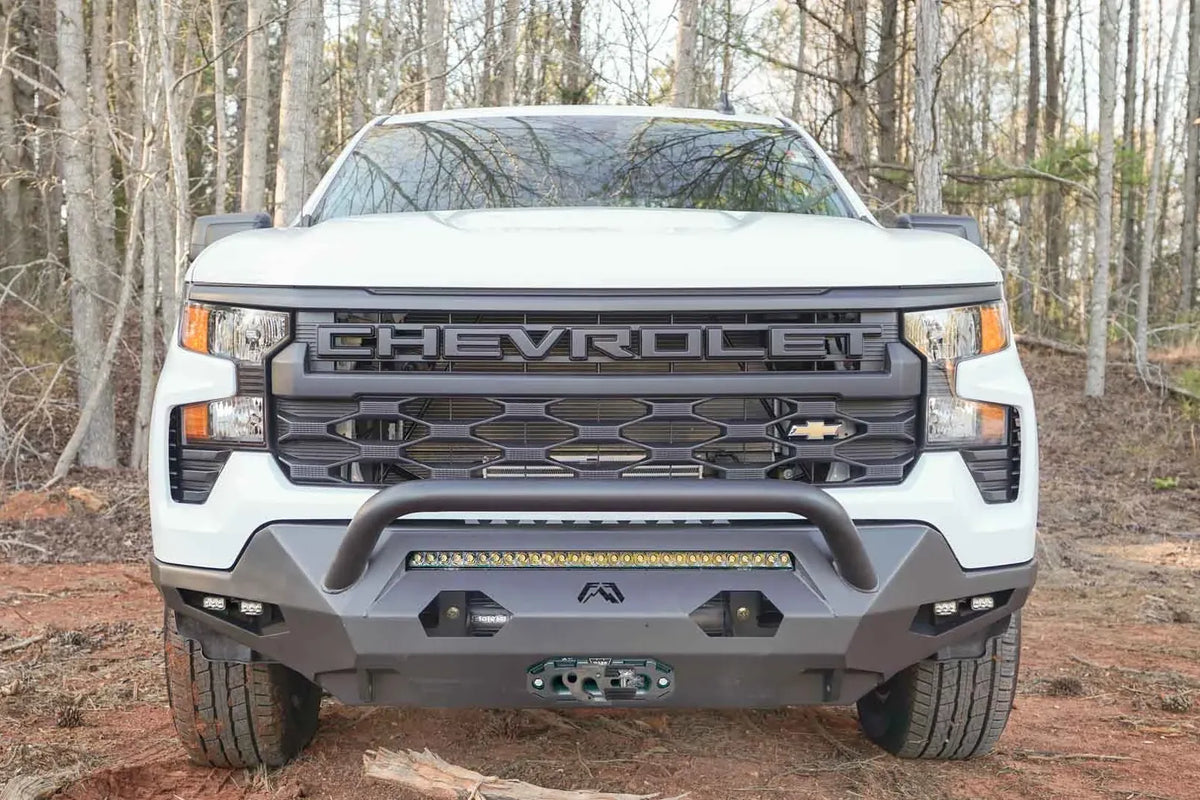 Fab Fours Matrix Front Bumper with pre-runner guard on a white Chevrolet Silverado 1500 outdoors on a dirt trail, front view with light bar, cube lights and winch fairlead