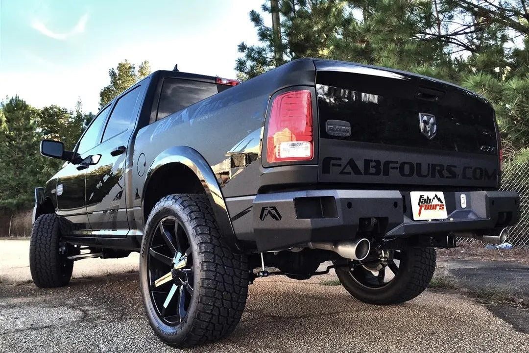 Fab Fours Premium rear bumper on a black Ram 1500, rear driver-side three-quarter view outdoors, showing the logo end caps, plate recess, and exhaust cutout