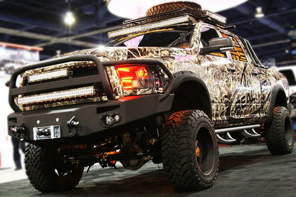 Fab Fours Premium front bumper with full guard on a camo-wrapped Ram 2500, front driver-side three-quarter view at an indoor show, showing the grille guard with light bar mounts, round fog lights, and winch