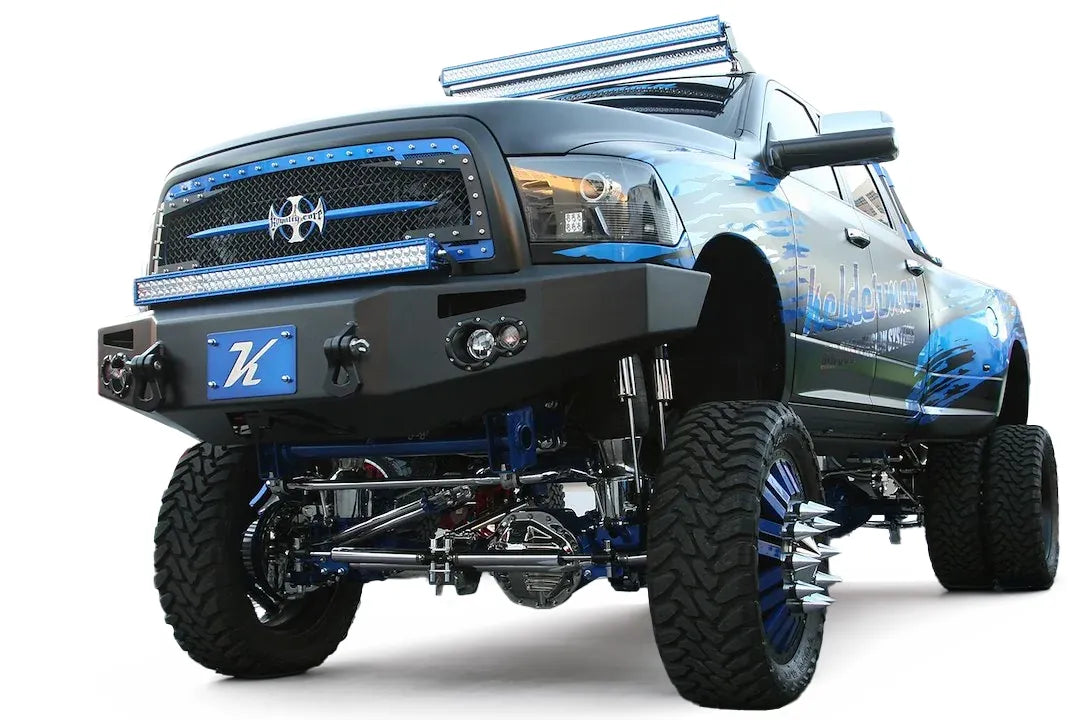 Fab Fours Premium front bumper on a black and blue Ram 2500, front driver-side three-quarter view, showing the plated winch bumper face, round fog lights, and shackle mounts