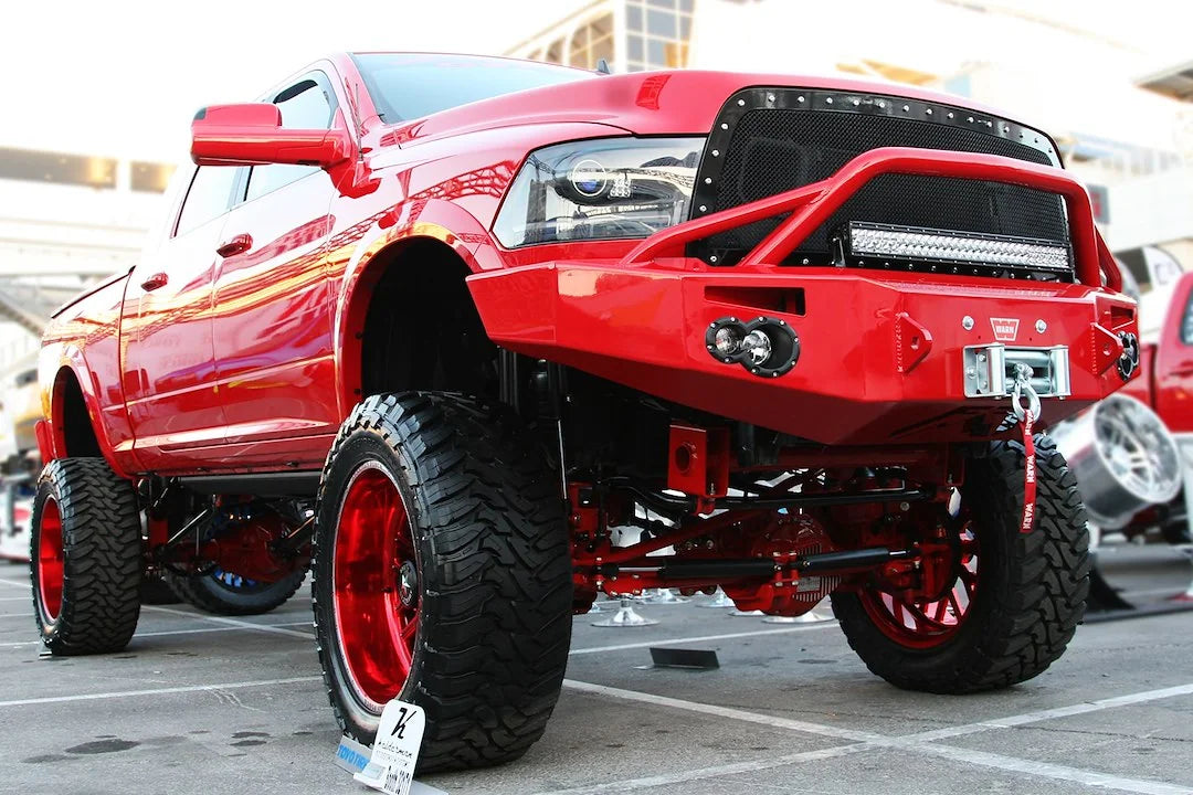 Fab Fours Premium front bumper with pre-runner guard in custom red on a red Ram 2500, front driver-side three-quarter view outdoors, showing the pre-runner hoop, winch with hook, and round fog lights