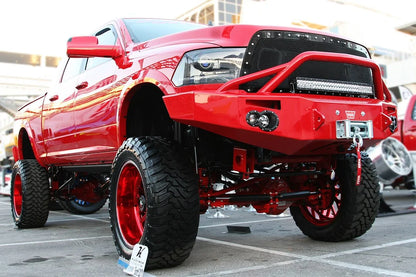 Fab Fours Premium front bumper with pre-runner guard in custom red on a red Ram 2500, front driver-side three-quarter view outdoors, showing the pre-runner hoop, winch with hook, and round fog lights
