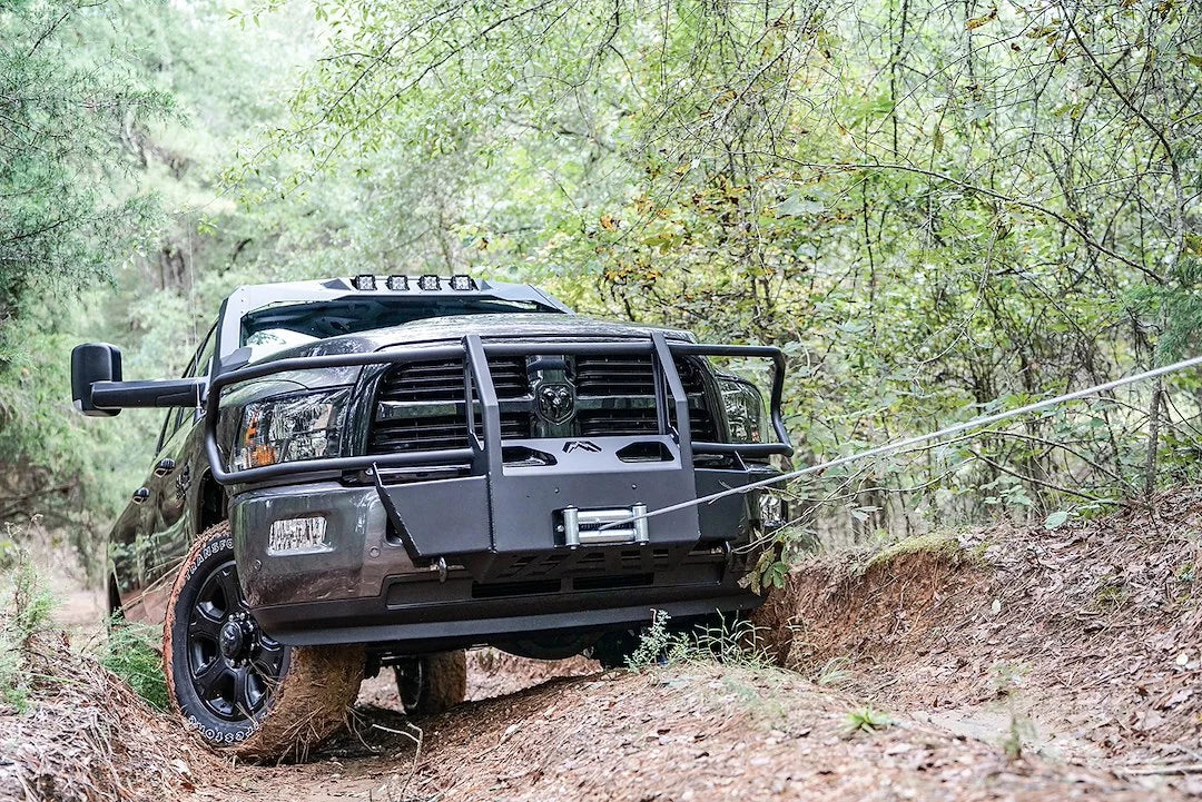 Fab Fours winch mount with full guard on a gray Ram 2500, front driver-side three-quarter view outdoors on a trail, showing the full grille guard, winch with line deployed, and headlight hoops