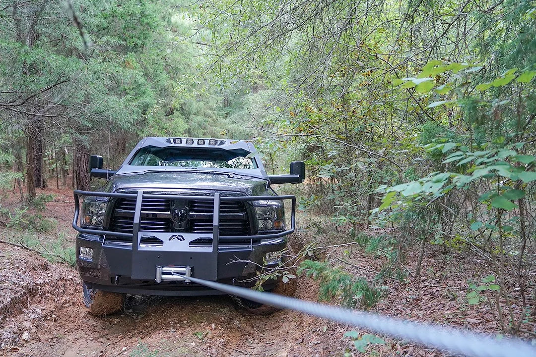 Fab Fours winch mount with full guard on a gray Ram 2500, straight-on front view outdoors in the woods, showing the full grille guard, winch spooling line, and plated mount face