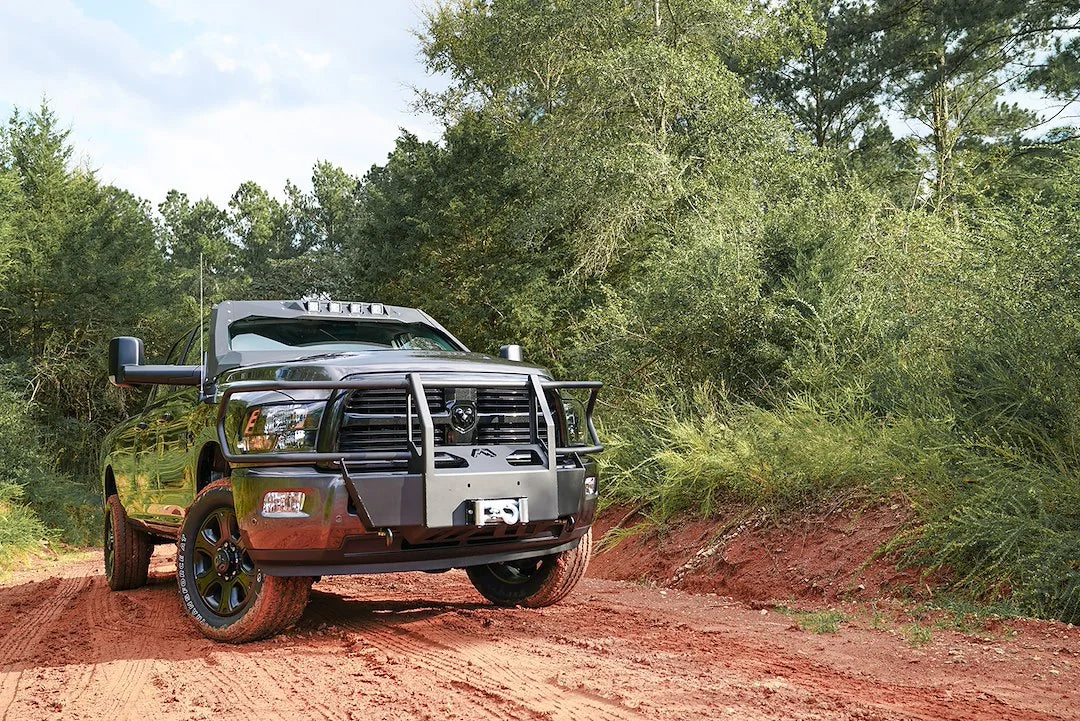 Fab Fours winch mount with full guard on a gray Ram 2500, front passenger-side three-quarter view outdoors on a dirt trail, showing the full grille guard, headlight hoops, and center winch