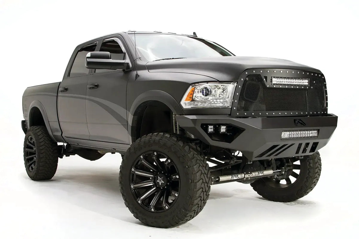 Fab Fours Vengeance front bumper on a gray Ram 2500, front passenger-side three-quarter view, showing the angular plated face, cube light pods, and slotted skid plate