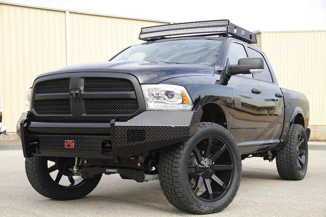 Fab Fours Black Steel front bumper on a black Ram 1500, front driver-side three-quarter view outdoors, showing the diamond-plate wings, mesh inserts and red logo badge