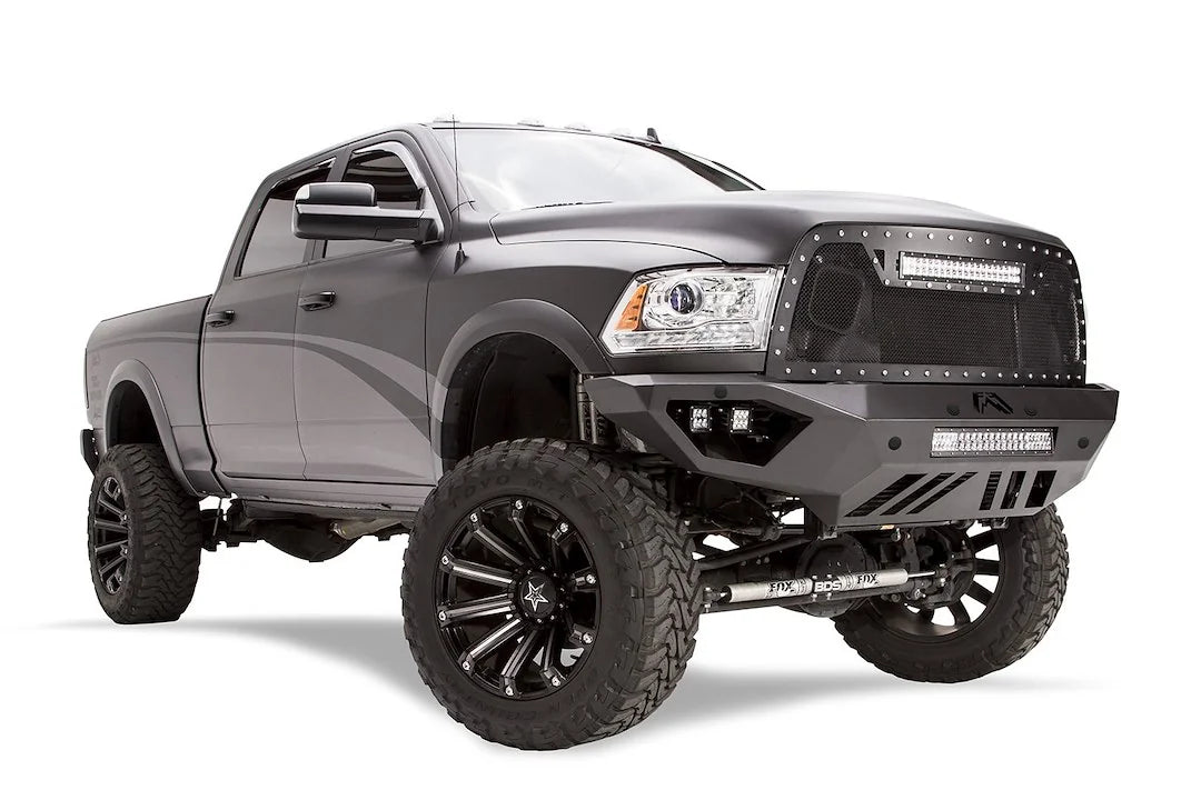 Fab Fours Vengeance front bumper on a dark gray lifted Ram 2500, front passenger-side three-quarter view, showing the integrated light bar, cube light pods and skid plate