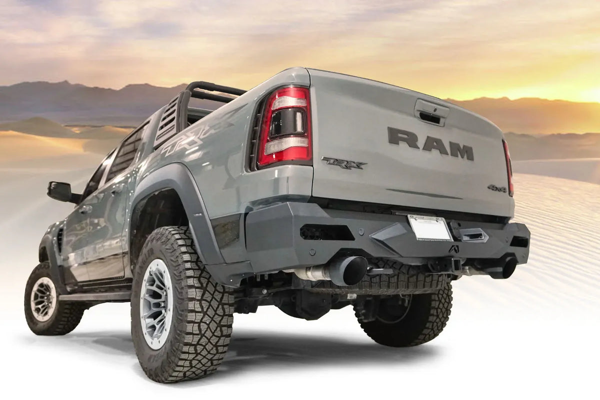 Fab Fours Vengeance rear bumper on a gray Ram 1500 TRX with sport bar, driver-side rear three-quarter view with desert sunset background, with parking sensors and hitch receiver