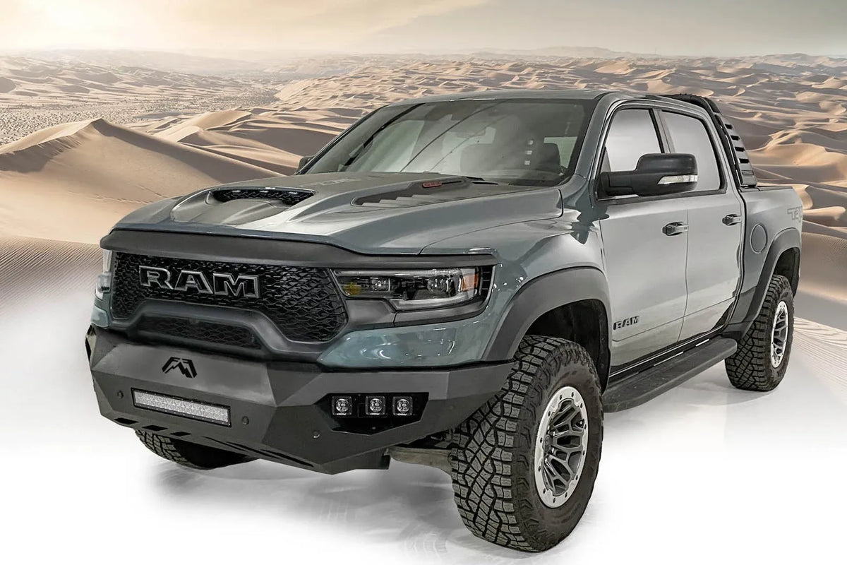 Fab Fours Vengeance front bumper on a gray Ram 1500 TRX, front driver-side three-quarter view against a desert backdrop, showing the low-profile bumper, light bar and wraparound end caps