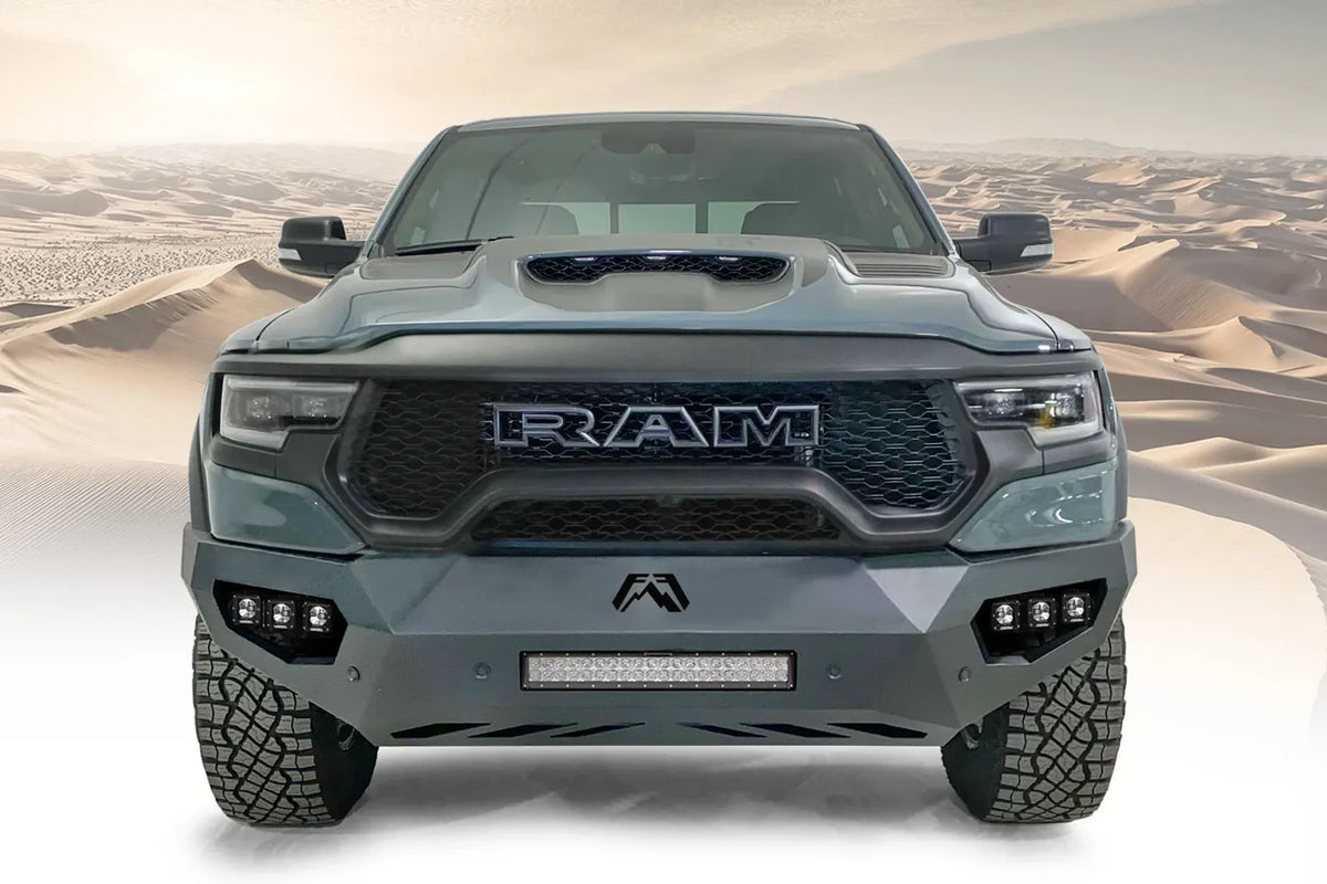 Fab Fours Vengeance front bumper on a gray Ram 1500 TRX, straight-on front view against a desert backdrop, showing the sculpted bumper face, center light bar and cube light pods