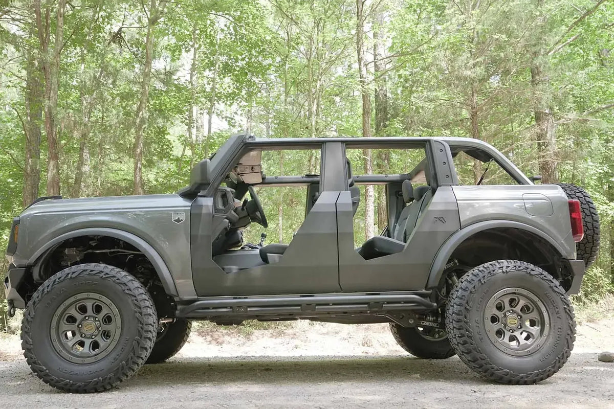 Fab Fours Trail Doors on a lifted gray four-door Ford Bronco on a wooded trail, driver-side profile, with the angular steel doors and Fab Fours logo on the rear door