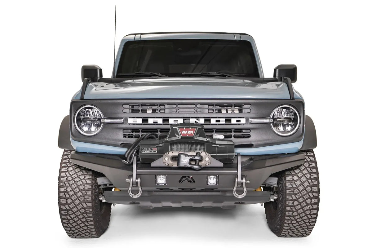 Fab Fours Stubby bumper on a blue-gray Ford Bronco, straight-on front studio view, showing the compact stubby bumper, winch mount, D-ring shackles and cube lights