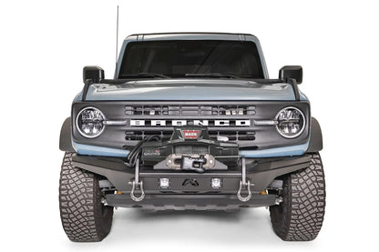 Fab Fours Stubby bumper on a blue-gray Ford Bronco, straight-on front studio view, showing the compact stubby bumper, winch mount, D-ring shackles and cube lights