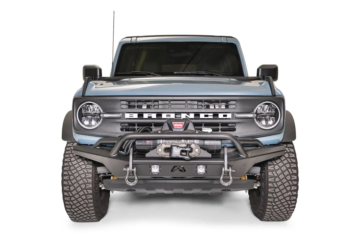 Fab Fours Stubby bumper with pre-runner guard on a blue-gray Ford Bronco, straight-on front studio view, showing the pre-runner hoop, winch mount, D-ring shackles and cube lights