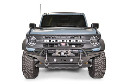 Fab Fours Stubby bumper with pre-runner guard on a blue-gray Ford Bronco, straight-on front studio view, showing the pre-runner hoop, winch mount, D-ring shackles and cube lights