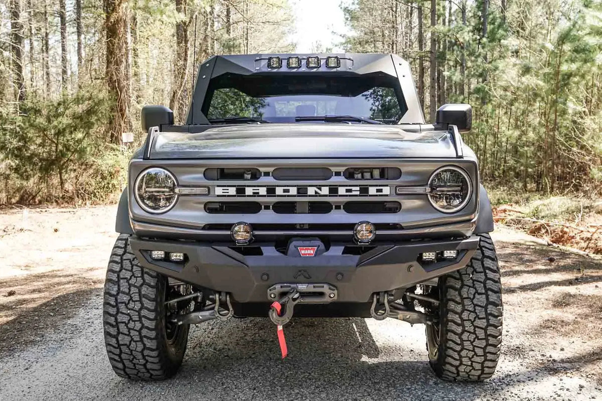 Fab Fours Lifestyle winch front bumper on a lifted gray Ford Bronco on a gravel road in the woods, front view, with Warn winch, pod lights, and D-ring shackles