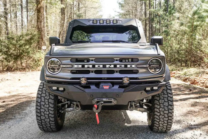 Fab Fours Lifestyle winch front bumper on a lifted gray Ford Bronco on a gravel road in the woods, front view, with Warn winch, pod lights, and D-ring shackles