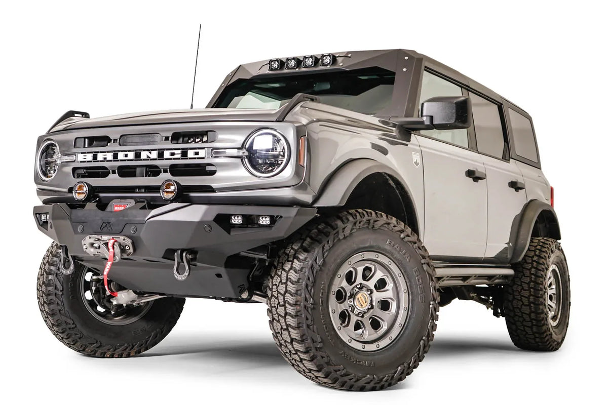 Fab Fours Lifestyle winch front bumper on a lifted gray Ford Bronco, driver-side front three-quarter view, with Warn winch, D-ring shackles, round pod lights, and cube lights