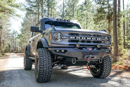 Fab Fours Lifestyle winch front bumper on a lifted gray Ford Bronco on a gravel road in the woods, passenger-side front three-quarter view, with Warn winch and D-ring shackles