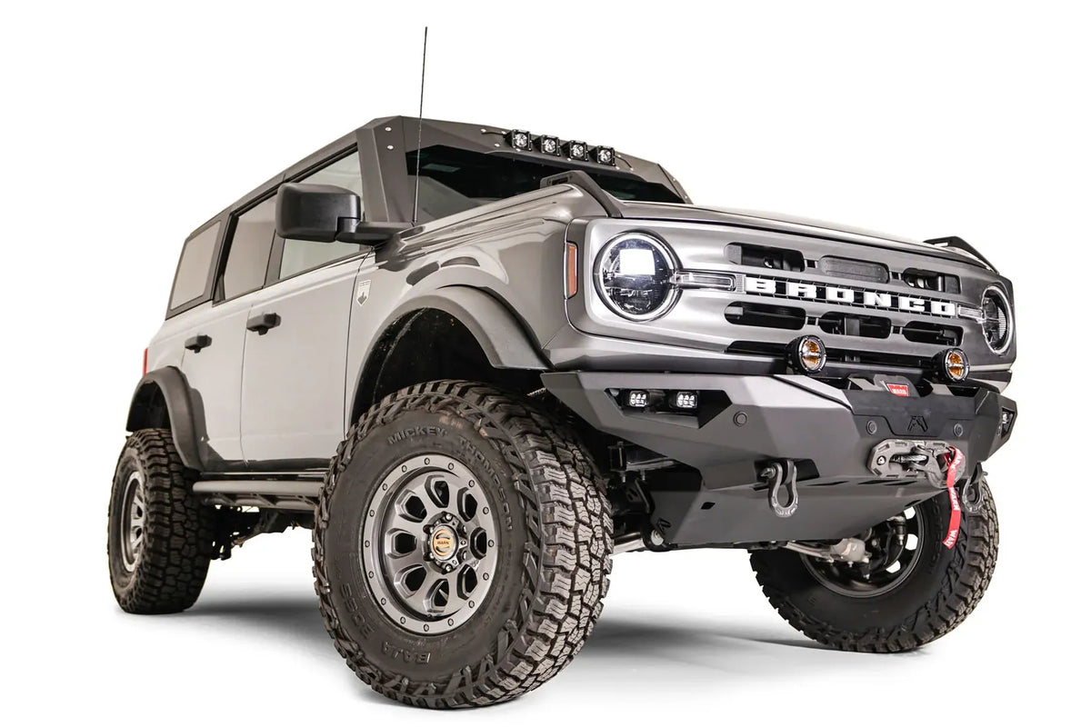 Fab Fours Lifestyle winch front bumper on a lifted gray Ford Bronco, passenger-side front three-quarter view, with Warn winch, D-ring shackles, round pod lights, and cube lights