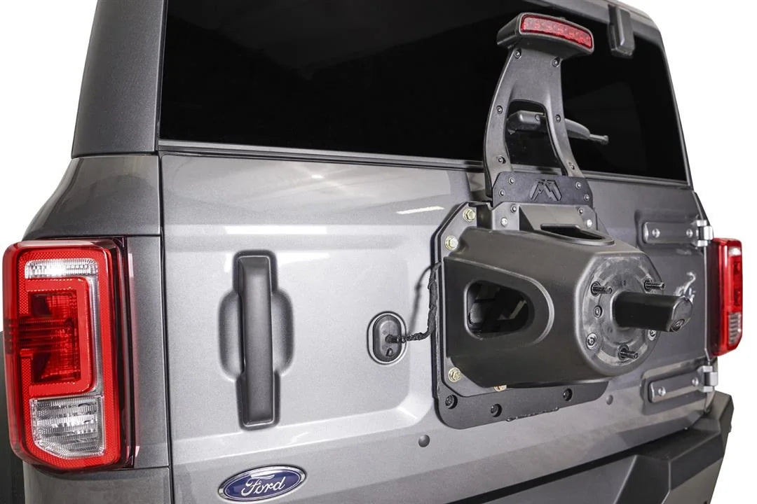 Close-up of the Fab Fours tire carrier relocation bracket on a Ford Bronco tailgate with the spare removed, showing the factory carrier and raised third brake light bolted to the steel plate