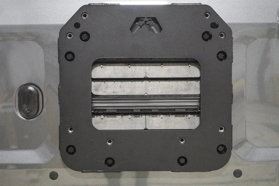 Close-up of the Fab Fours tire carrier relocation bracket bolted to a Ford Bronco tailgate, showing the matte black steel plate with Fab Fours logo cutout and mounting holes