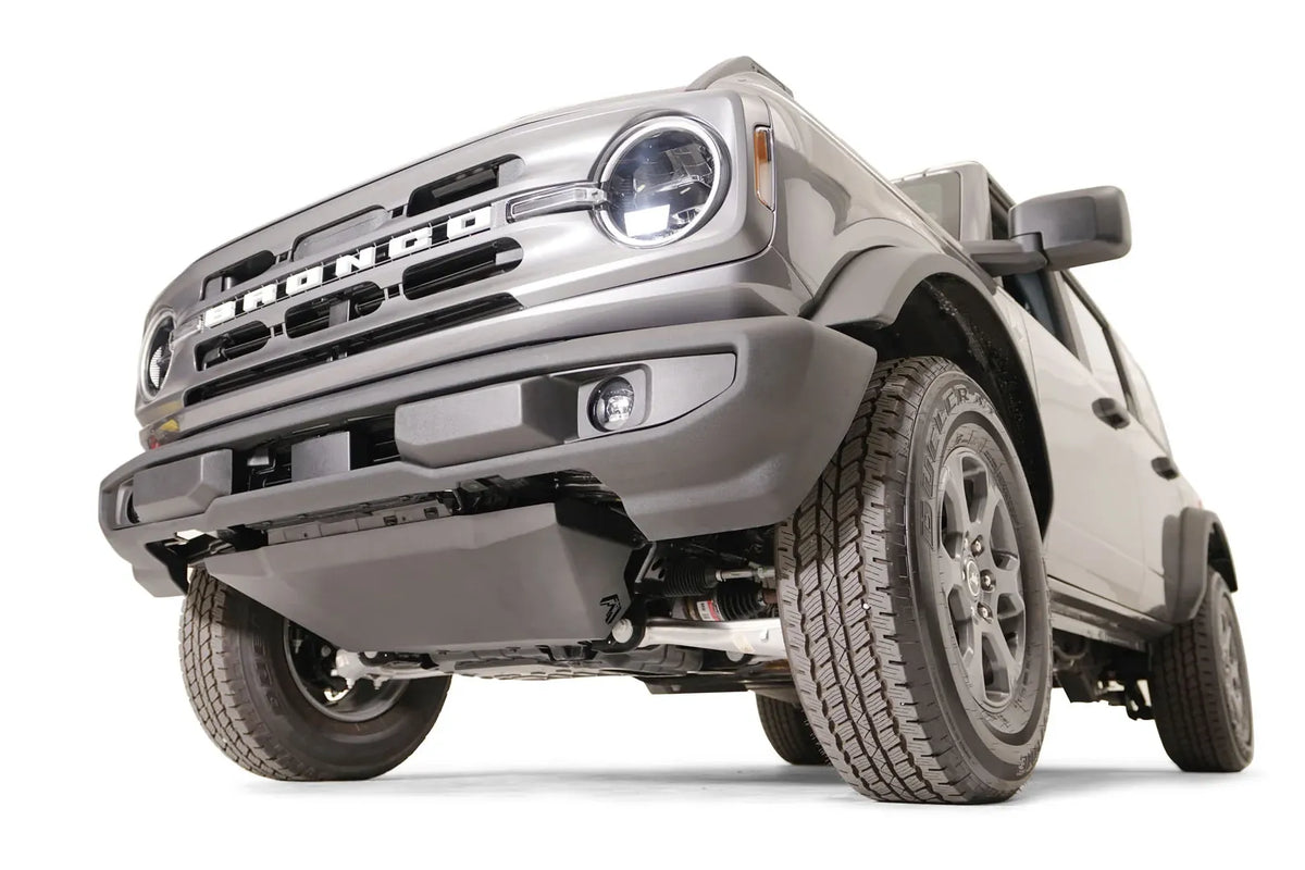 Fab Fours engine skid plate on a gray Ford Bronco with the factory front bumper, low front driver-side view, showing the black skid plate with the Fab Fours logo covering the engine below the bumper