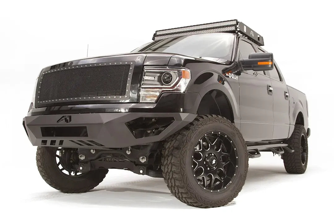 Fab Fours Vengeance front bumper on a black Ford F-150, front driver-side three-quarter studio view, showing the sculpted bumper face, angular end caps and lower skid section