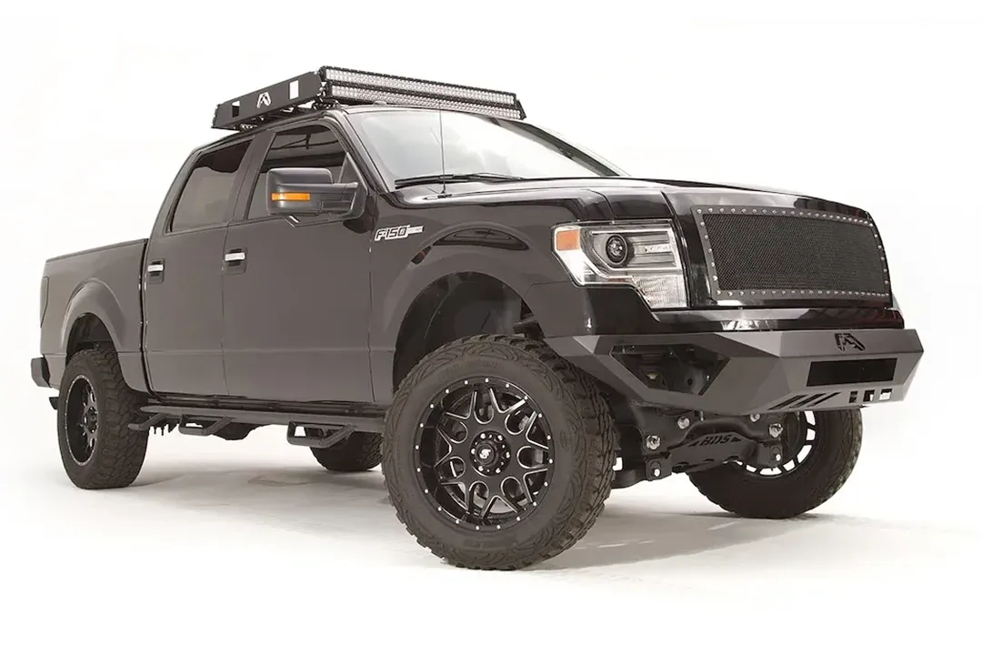 Fab Fours Vengeance front bumper on a black Ford F-150, front passenger-side three-quarter studio view, showing the low-profile bumper, end caps and center logo plate
