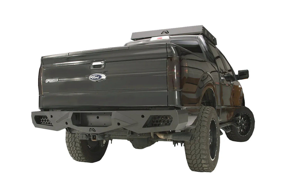 Fab Fours Vengeance rear bumper on a black Ford F-150, rear passenger-side three-quarter studio view, showing the sculpted rear bumper, honeycomb corner vents and center hitch opening