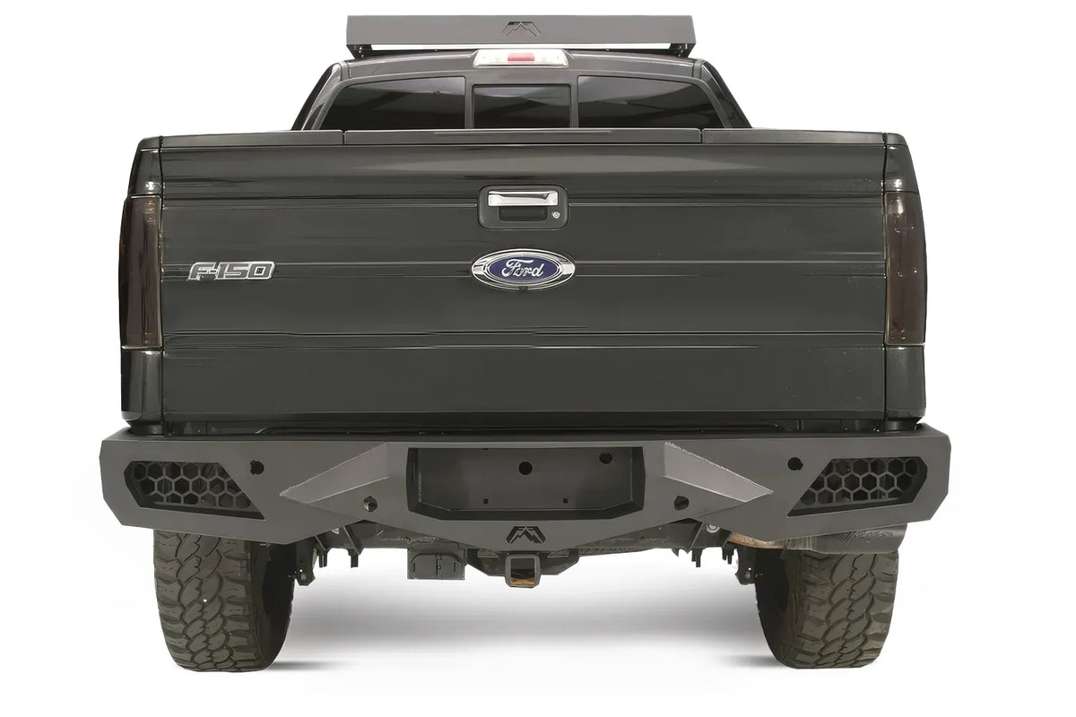 Fab Fours Vengeance rear bumper on a black Ford F-150, straight-on rear studio view, showing the sculpted bumper face, honeycomb corner vents and center hitch opening