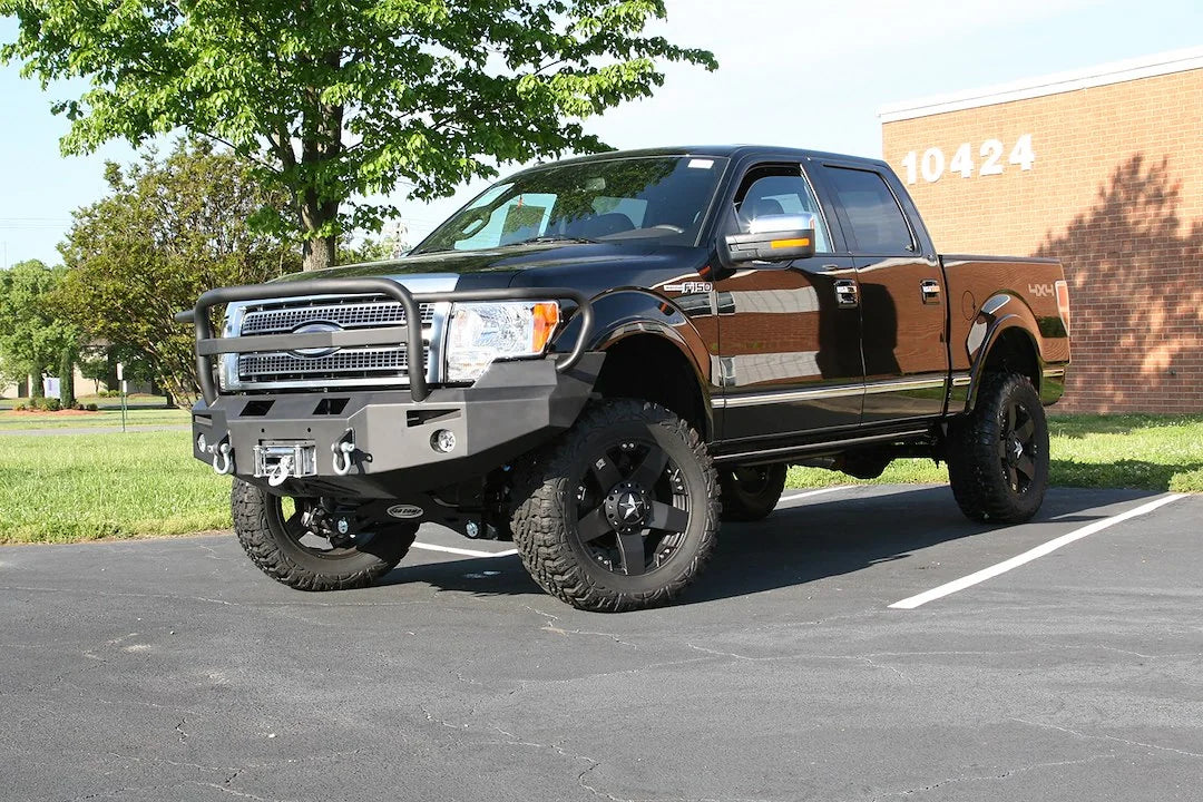 Fab Fours Premium Winch front bumper with full guard on a black Ford F-150, front driver-side three-quarter view outdoors, showing the full grille guard, bumper wing and winch-ready center section