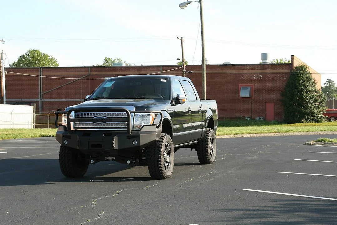 Fab Fours Premium Winch front bumper with full guard on a black Ford F-150, front view in a parking lot from a distance, showing the full grille guard and heavy-duty bumper