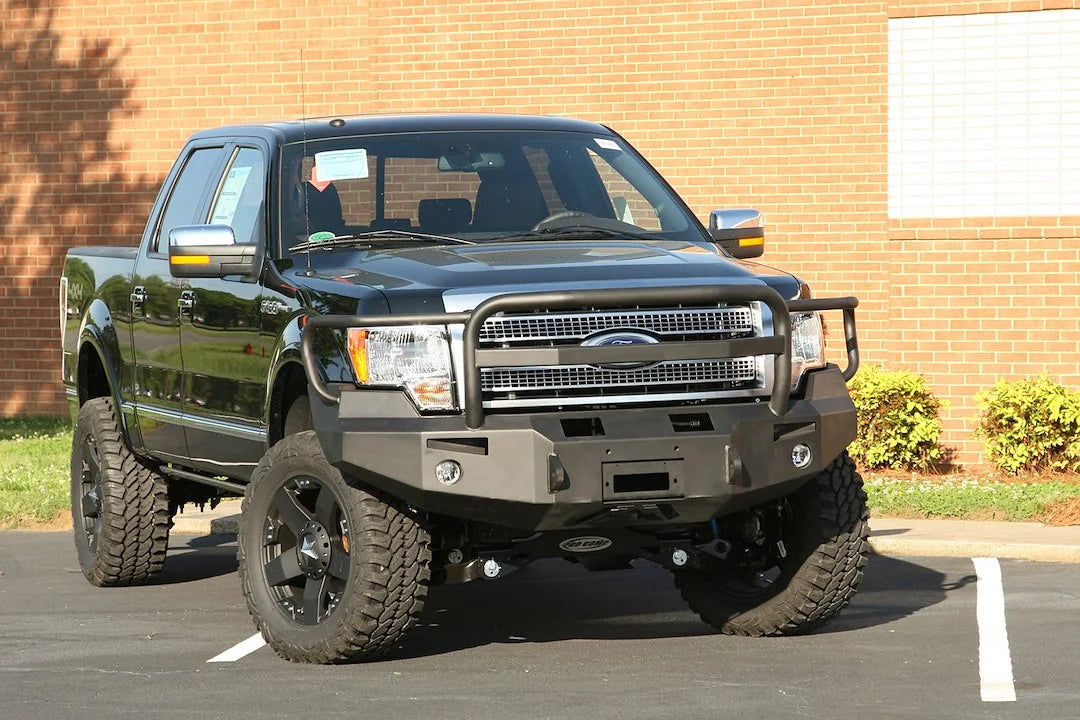 Fab Fours Premium Winch front bumper with full guard on a black Ford F-150, front passenger-side three-quarter view outdoors, showing the full grille guard, winch-ready center section and fog lights