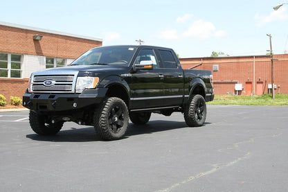 Fab Fours Premium Winch front bumper on a black Ford F-150, front driver-side three-quarter view in a parking lot, showing the heavy-duty bumper, winch-ready center section and fog lights