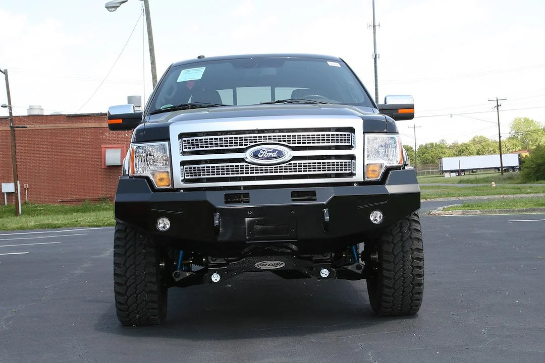 Fab Fours Premium Winch front bumper on a black Ford F-150, straight-on front view in a parking lot, showing the heavy-duty bumper face, winch opening and fog lights