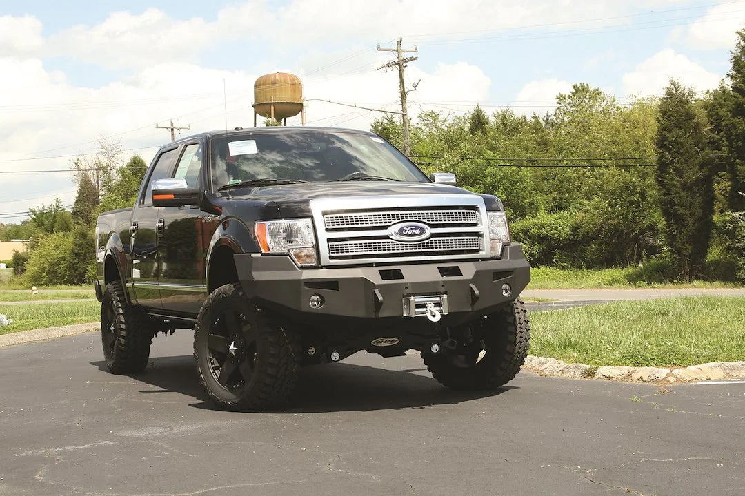 Fab Fours Premium Winch front bumper on a black Ford F-150, front passenger-side three-quarter view outdoors, showing the heavy-duty bumper, mounted winch and fog lights