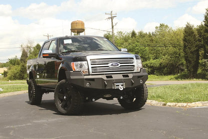 Fab Fours Premium Winch front bumper on a black Ford F-150, front passenger-side three-quarter view outdoors, showing the heavy-duty bumper, mounted winch and fog lights