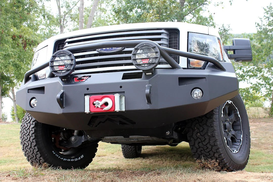 Fab Fours Premium Winch front bumper with pre-runner guard on a white truck, low close-up front view outdoors, showing the pre-runner hoop with mounted lights, winch fairlead and fog lights