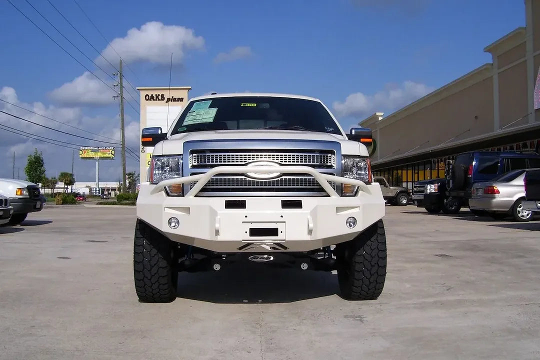 Fab Fours Premium Winch front bumper with pre-runner guard on a white Ford F-150, straight-on front view in a parking lot, showing the pre-runner hoop, heavy-duty bumper face and fog lights