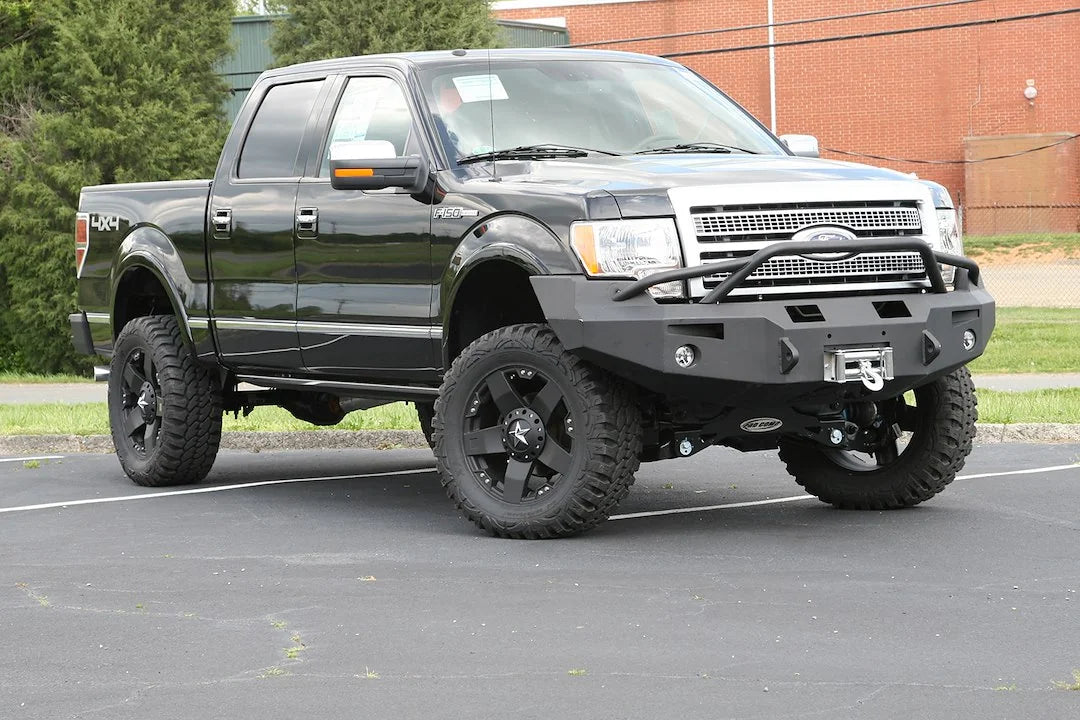 Fab Fours Premium Winch front bumper with pre-runner guard on a black Ford F-150, front passenger-side three-quarter view outdoors, showing the pre-runner hoop, mounted winch and fog lights